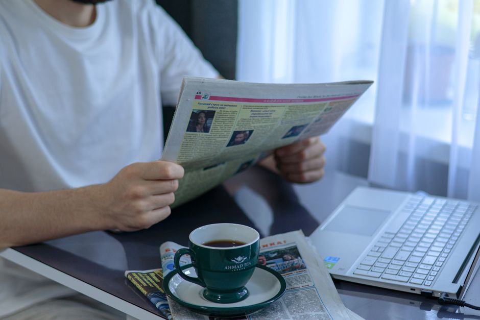 Person med kaffe och tidning vid ett skrivbord med laptop i dagsljus