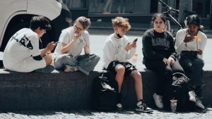 Slang från orten – ordlista med de vanligaste orden och vad de betyder Grupp ungdomar sitter på en trottoar med mobiler och pratar, illustration till artikel om orten slang